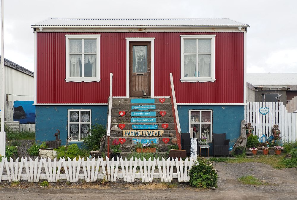 The house has two storeys. The front steps in the center of the house lead up to the front door at the upper floor, with a window on either side of it. The upper storey is painted red. Below it, the lower storey appears to be partially under ground. One window shows on either side of the central stairway. This floor is painted blue. The trim on the windows and door is painted white. The stairs have, in the middle of each step, something painted in white on blue in Icelandic. The words are framed on either side of every second step with a red heart. The small yard is edged with a white picket fence.