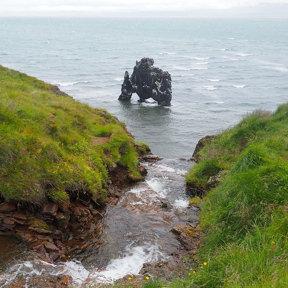 Seen from uphill at a waterfall, the water flows straight ahead down to the sea, grassy hills on either side of the stream. In the sea off the shore is a rock formation standing on three legs.