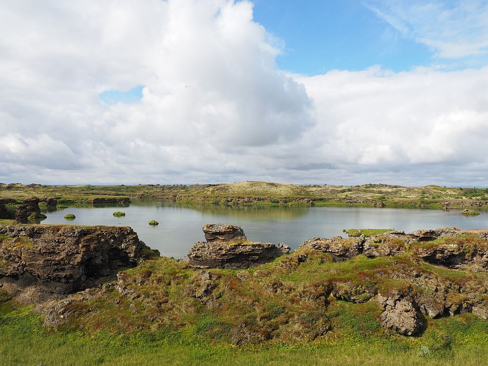 A still lake, edged by brownish rock and some low grassy plants.