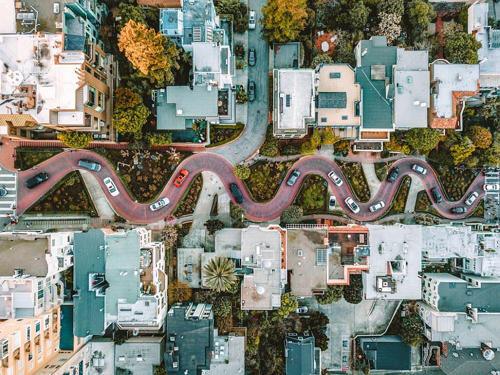 A drone view straight down of Lombard Street. The eight curves of the street are visible, with cars on the street. On either sides, the mostly flat roofs of the buildings on either side, with lots of greenery along the road and between and behind the buildings.