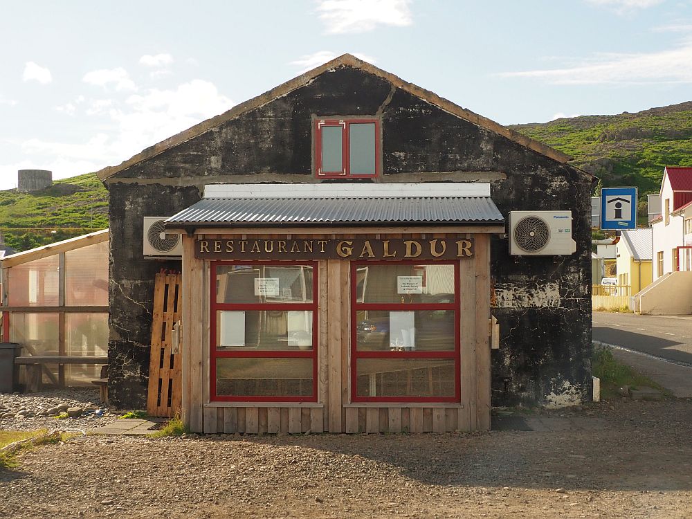 A brown, not recently painted, low building of one story. Across the front are some big windows and the words "Restaurant Galdur". 