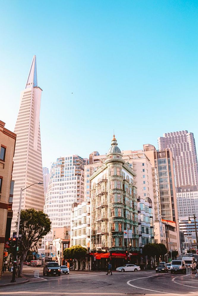 In the center, the Sentinal building is triangular, its sharpest point at the intersection of two streets. Traffic waits at its base while pedestrians cross. To the left of the picture is the Transamerica pyramid in the distance, along with a jumble of less distinctive tall buildings.