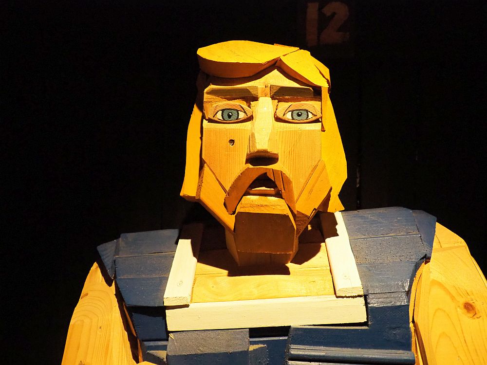 A human figure made of wood. Here only photographed from the chest up. It is a man with blond hair and a mustache and beard. The figure is expressed in rough-edged shapes of wood in different colors, almost impressionistic.