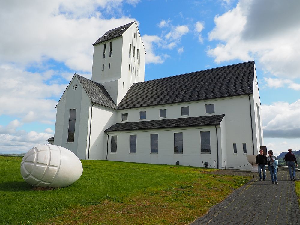 The church is white with a square tower and a grey roof, here seen from the side. On the grass next to it is a large egg, also white, with some ridges carved into one end. The egg is huge, probably about a meter across. 3-week Iceland itinerary