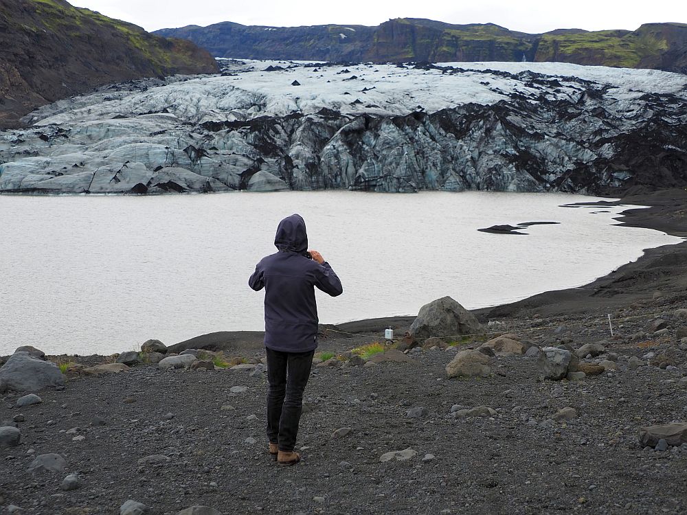 In the foreground, Albert stands with his back to the camera looking toward the glacier, holding his phone to take a picture. In front of him is the meltwater lake, and beyond that is the glacier: a feild of bluish ice with an almost vertical shelf at the lakeside where chunks have broken off.