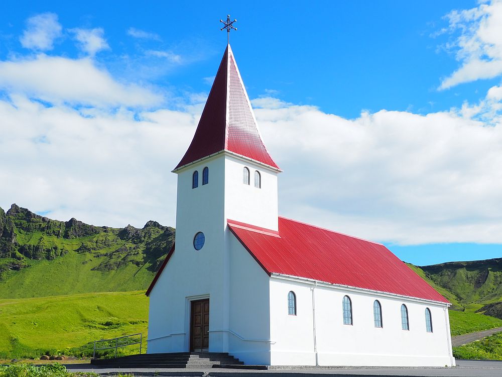 The church is simple with plain white sides and a bright red roof on the church and the steeple. The steeple is square with a few small windows near the top. The windows along the sides of the church are arched at their to ends.