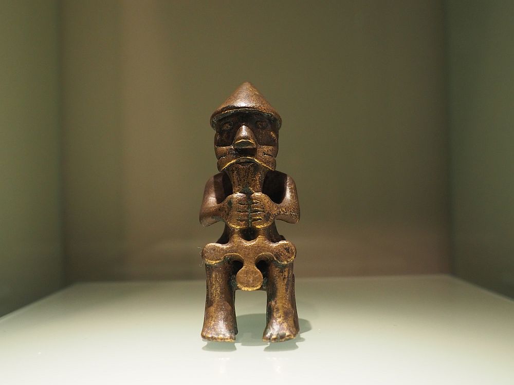 The figure is in a sitting position facing the camera. It wears a pointy hat, has a mustache, and holds the hammer or cross in both hands in front. It reaches from his chin to his knees.