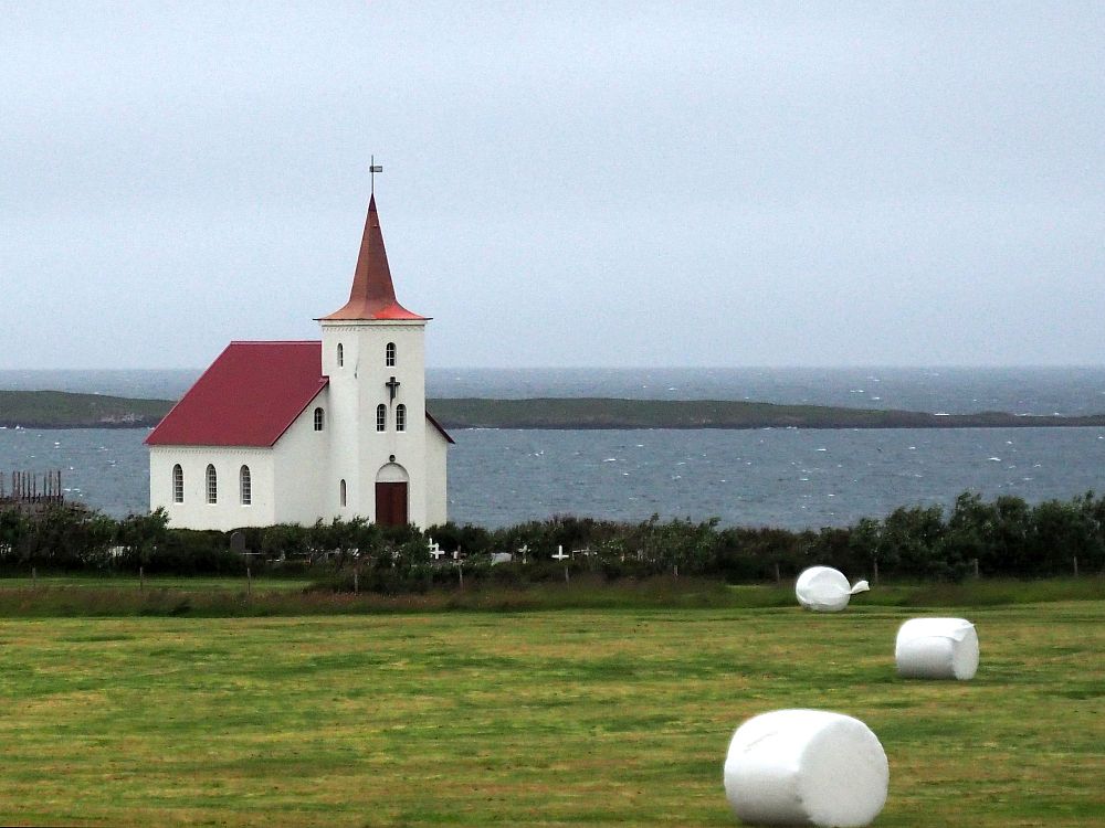 A simple small church: white walls, red roof, simple pointy steeple, faces away from the sea behind it. In front is a field with bales of hay scattered on it, all wrapped in white plastic.