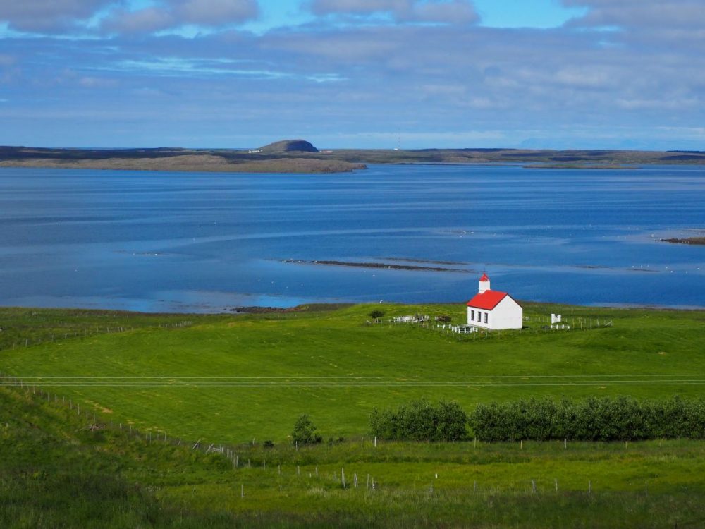 A green field in the foreground, blue sea beyond, with some low land on the other side of a bay. In the field is a white church with a red roof facing the sea. Passed somewhere along the way on our 3-week Iceland itinerary.