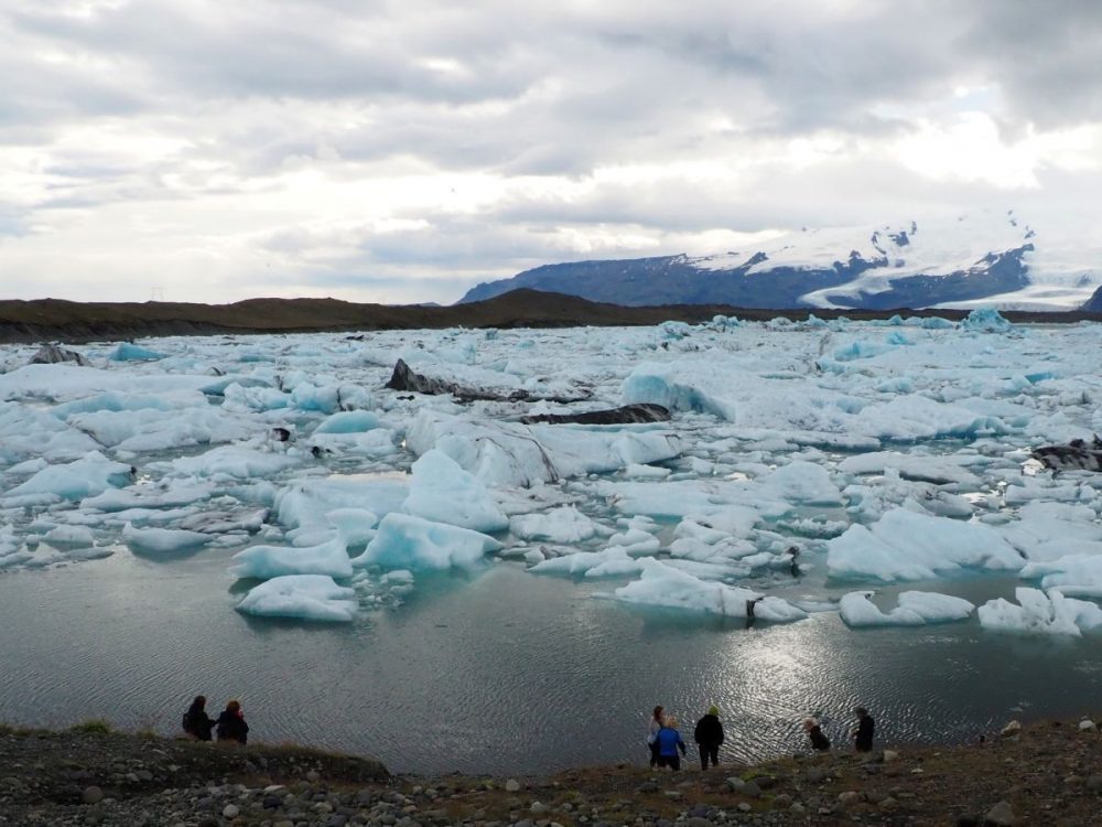 The lagoon is filled with chunks of ice, all looking bluish. Some open water is visible in the foreground, with a few people in sillhouette at the bottom of the picture. Behond the lagoon is a mountain covered in snow.