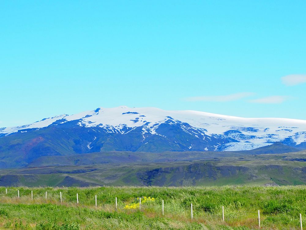 In the foreground, a green field, with a low fence marking its edge. Beyond, a mountain, covered in snow, against a blue sky.