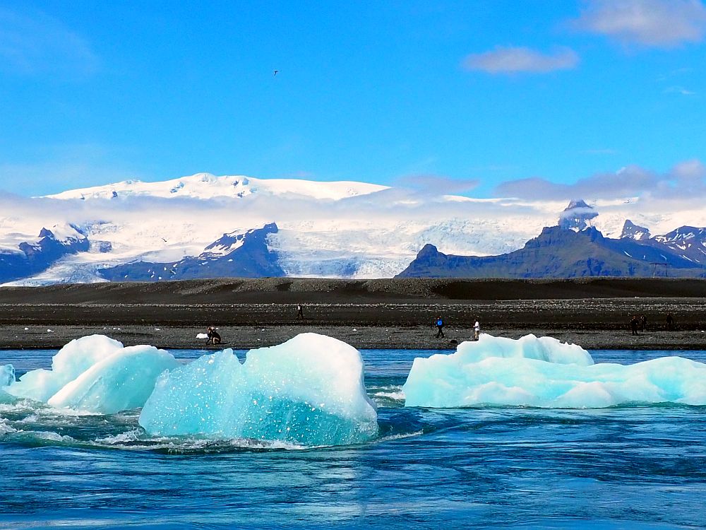 Blue chunks of ice float (or stand on the bottom) on darker blue water. The opposite bank of the river is black stone and a few people stand on the riverbank. Beyond that, the mountains rise, covered with snow and some clouds low over them. A glacier can be seen at the left of the photo, looking like a white river coming down the mountain.