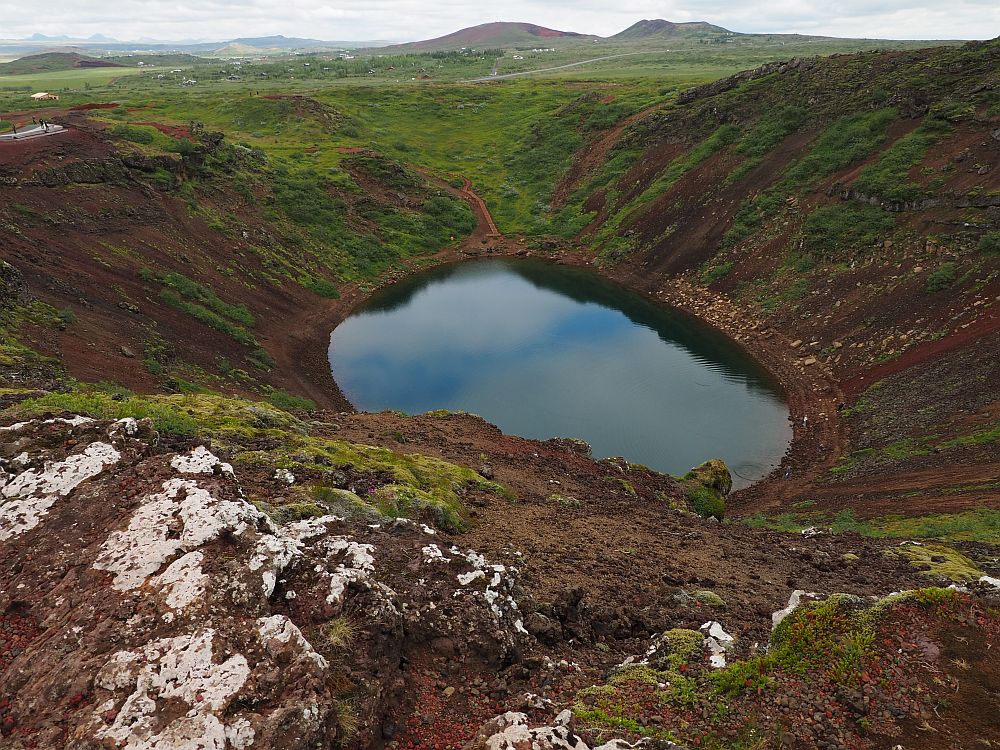 Looking down into the crater, the sides are brown with some low green cover, probably moss. The water is quite dark, reflecting the sky of white clouds and a bit of blue.