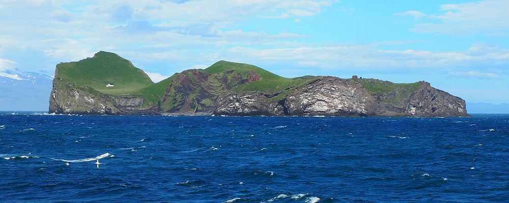 In the foreground, very blue sea. On the horizon, an island, edged by steep cliffs, green on their top. A single house is visible at one end.