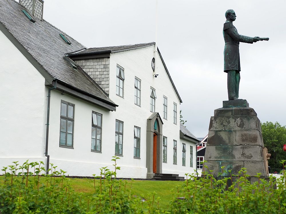 Seen from the side, looking along the front, the house is white with a grey roof and remarkably plain. Only two stories, it appears to be relatively small too. A statue stands in front of the house of a man, holding out a document in front of him.