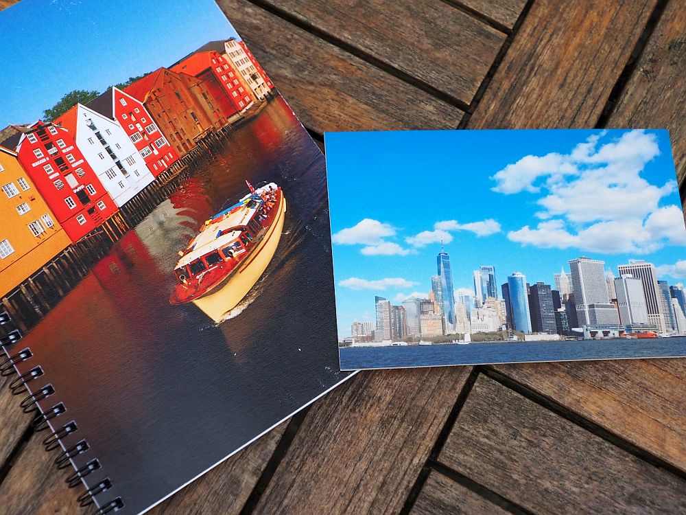 On the left, a spiral notebook with an image of brightly colored warehouses on a canal in Norway, a small boat passing in front of them. On the right, a photo of the New York City skyline against a blue sky with fluffy clouds. 