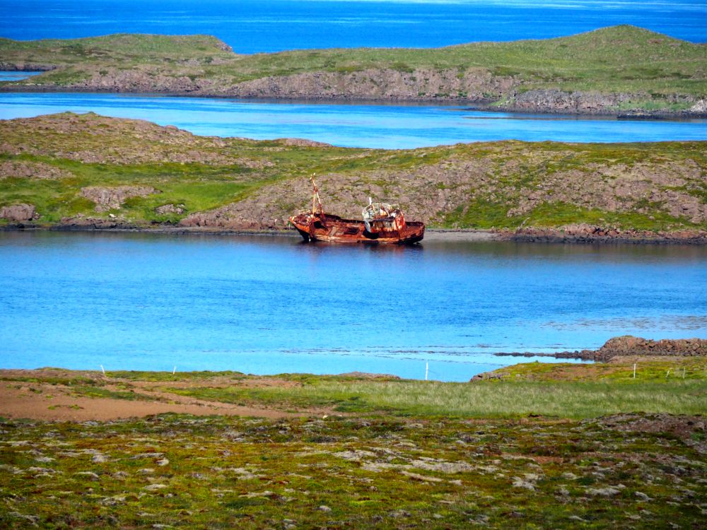 Across a body of water are some islands. Between the shore and the first island is the wreck of a fishing boat. It leans to the side, covered in rust. The water is bright blue here.