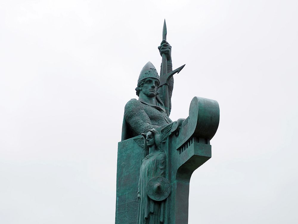 The man is an idealized Viking image: He holds a spear in his left hand pointing straight up. He has a pointed helmet. In front of him is a dragon figure, presumably to suggest a Viking ship. The statue is greenish.