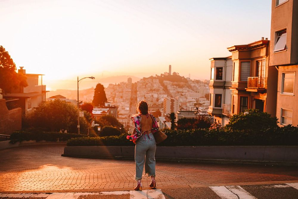 A sunset shot looking at the top curve of Lombard street, with a woman facing down the hill, her back to the camera. A few of the buildings on Lombard street are visible on either side, and in the center distance is a hill with Coit tower showing in sillhouette from the top against an orangy-yellow sky. 