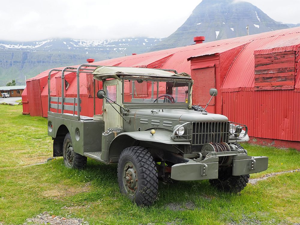 Front and center: a jeep or possibly a rover; it's hard to tell. It is khaki green and has an open back with seats along each side. Behind: a long red building of corrugated metal with a rounded roof: a quonset hut.