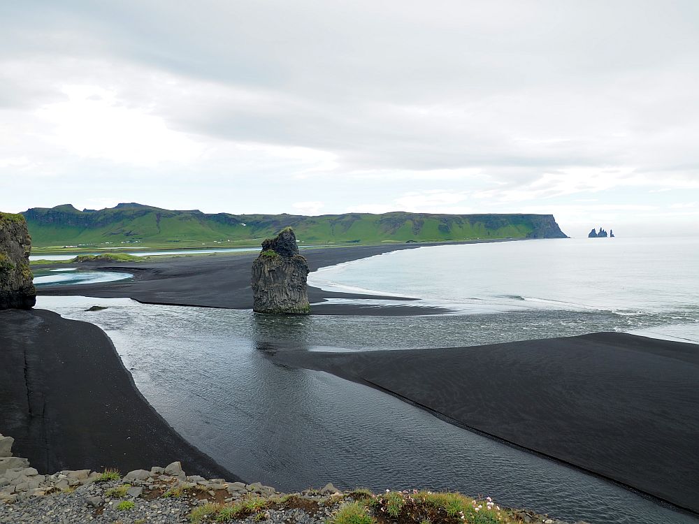 This view along the coast shows a wide black sandy beach, with a shallow flow of water crossing it to the sea at the right. A large rock formation stands on the beach, and some more a visible in the distance.