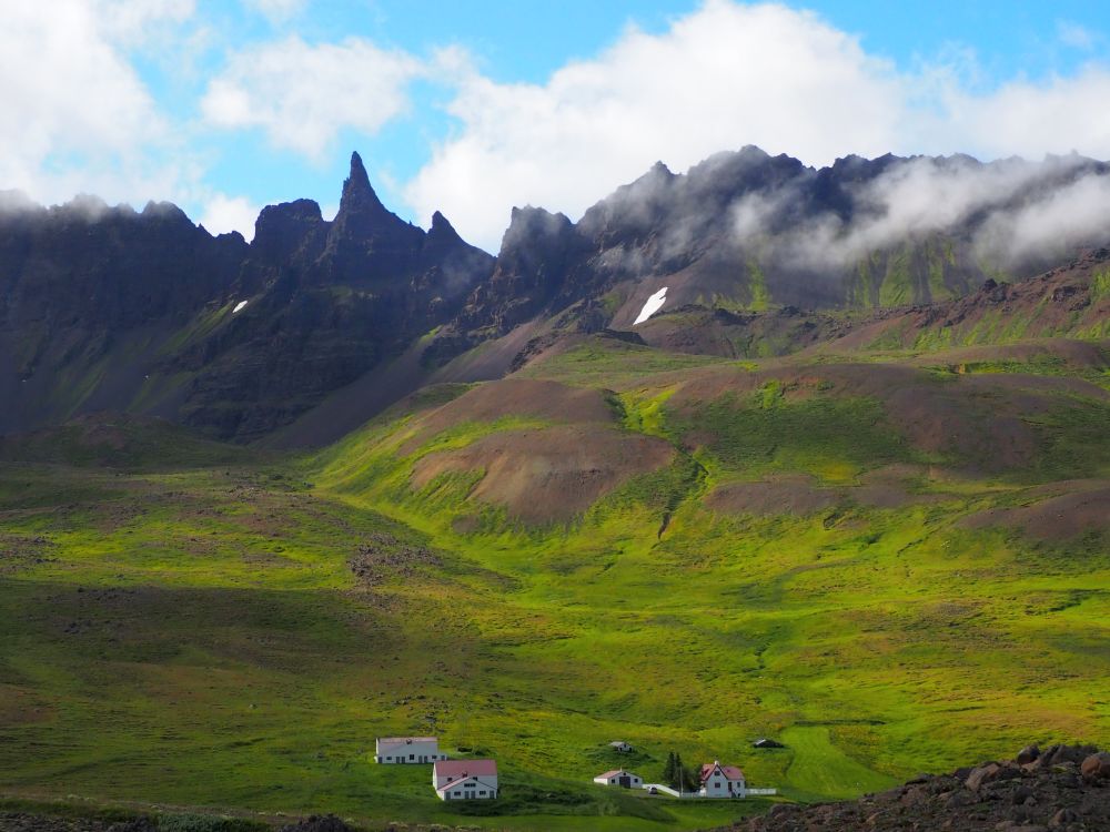 At the bottom of the photo is a cluster of houses, all of them painted white with red roofs. Looming behind them is a range of craggy mountains. Between the houses and the mountains the land rises but is grass-covered.