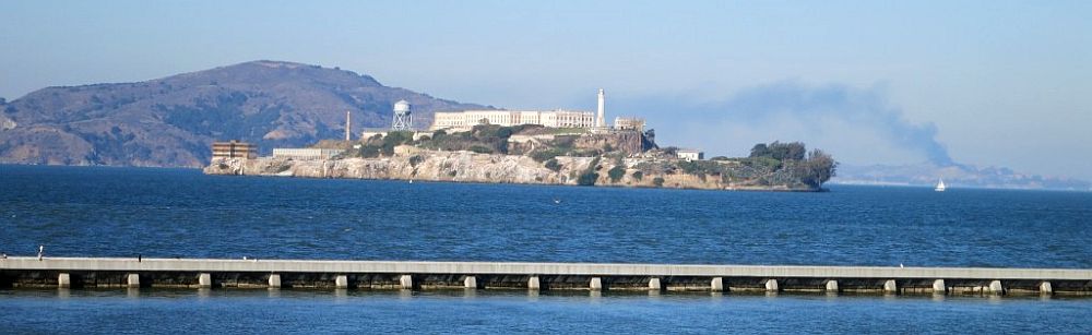 Across blue water, the island of Alcatraz looks like it's topped by a castle. Beyond that, further across the water, a small mountain, and a plume of smoke even further in the distance.