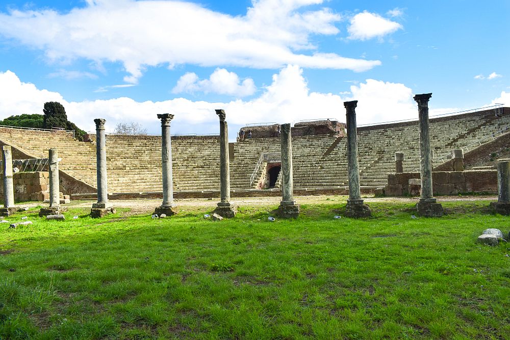 In the background, a half-circle of stone seats form an ampitheater. In the foreground, a row of columns, some partly ruined.