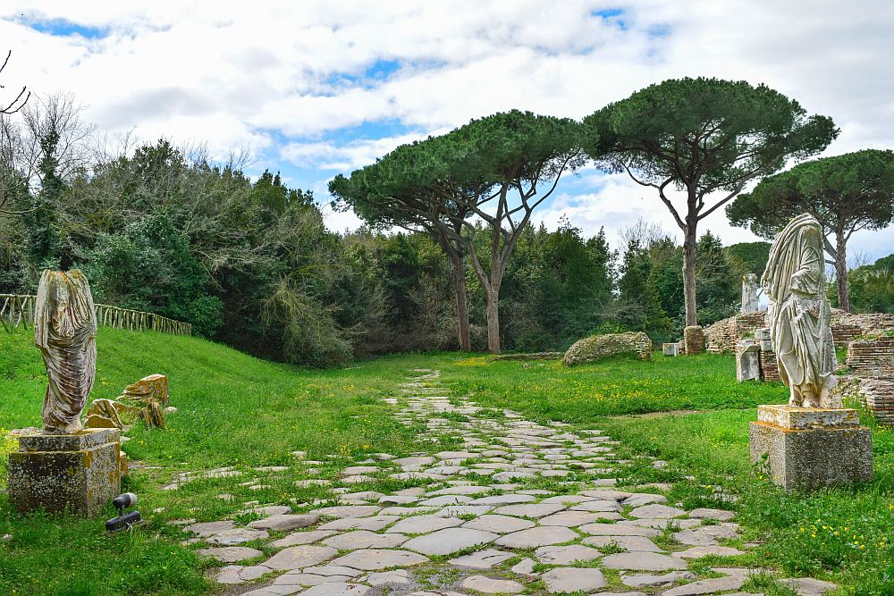 A path is paved in flat stones of various shapes and extends straight ahead into a grassy space. Beyond that it disappears into trees. On the left and the right are two human statues on pedestals. Both are dressed in robes but their heads are missing.