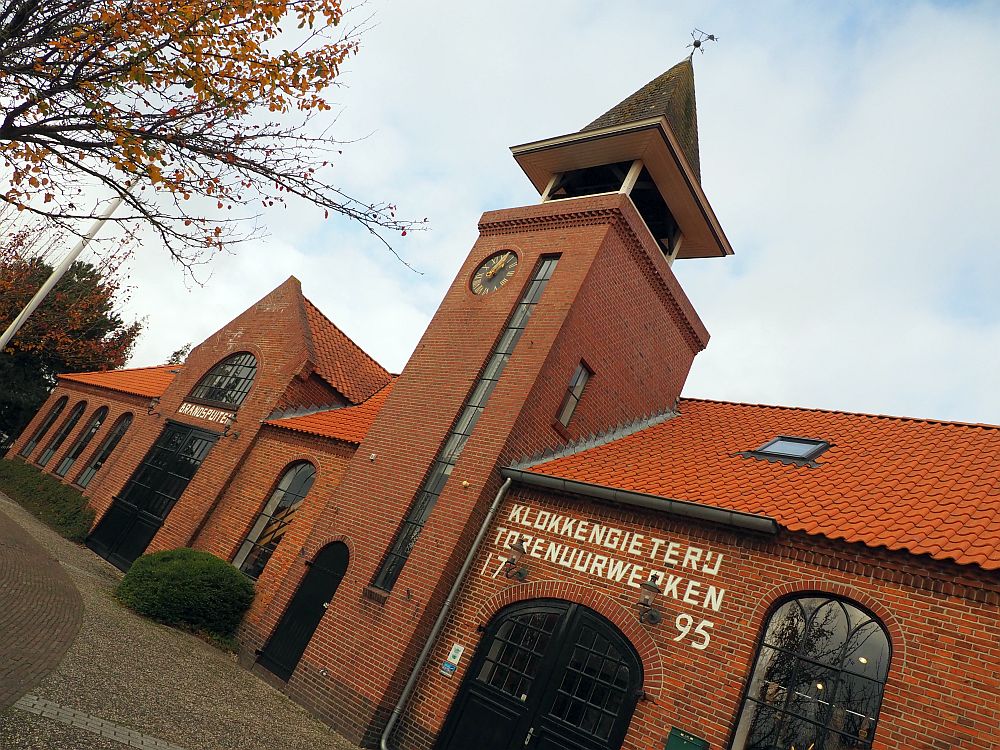 The photo, taken at an angle, looks along the front of the Bell Foundry museum building. It is red brick, with arched windows and doorways, just one storey except for a small tower which looks to be two or three stories and has a small pointed roof on top.