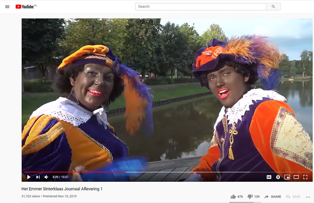 Two Zwarte Piets (Black Petes) smile at the camera. Both wear the typical bright Moorish costume and a beret with a feather. Both are in dark brown facepaint and have bright red lipstick on. 