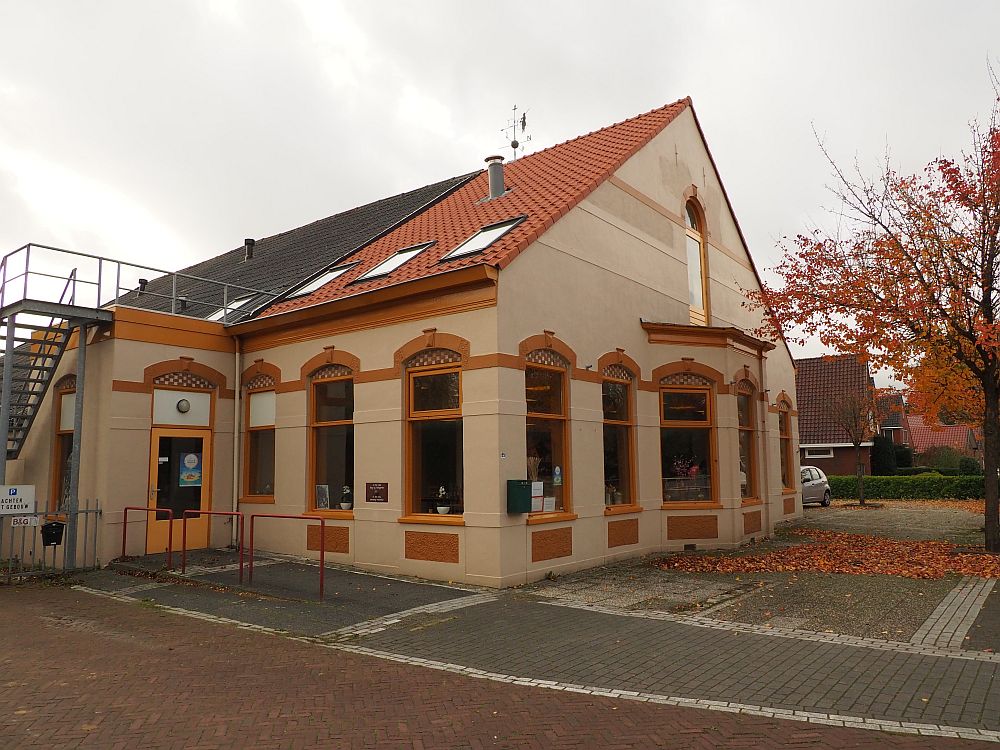 A former farmhouse, the front has elegant windows on the ground floor and one central window on the upper floor under the red slanted roof. In this view from an angle, some of the back end of the building can be seen: what would have once been the barn. It has a grey roof. 