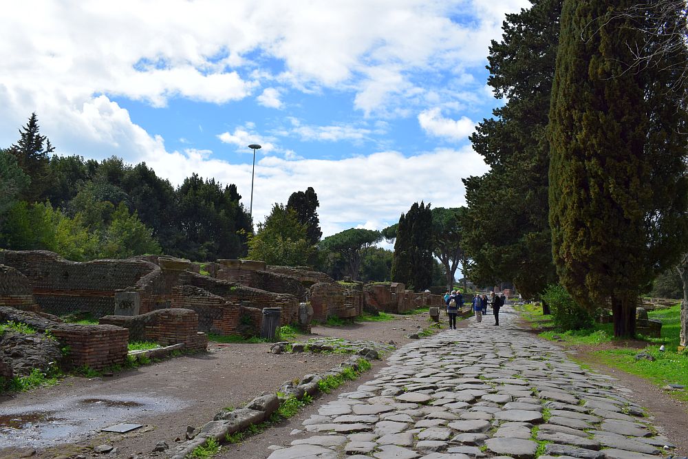 A wide road, about the width of a modern single lane, stretches ahead into the distance, trees on the right along its side and low stone ruins of buildings on the left, reduced to low walls. The road is paved with flat stones of various shapes.