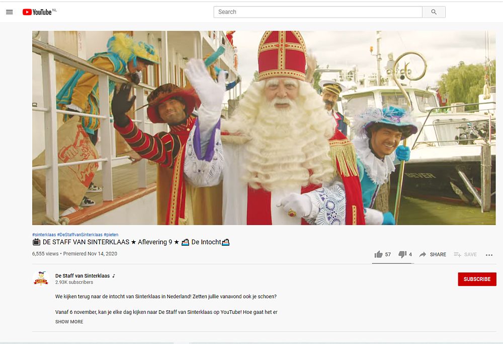 Sinterklaas faces the camera, waving his hand. He has a long beard and a red and gold mitre on his head. He wears a red and gold robe and white gloves. Two Petes are visible behind him to the left and right. Both are dressed in colorful outfits. Both have their natural white skin. The one on the left waves at the camera. The one of the right smiles. 