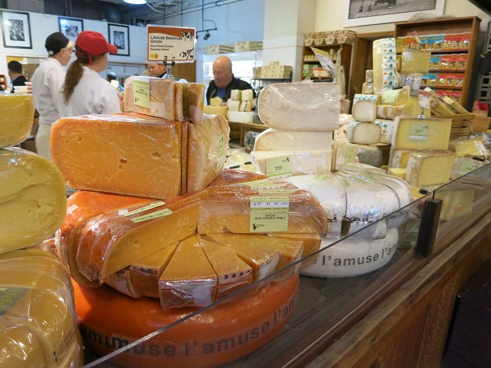 Various cheeses on display in the foreground. Behind, two people in white shirts speak to a customer.