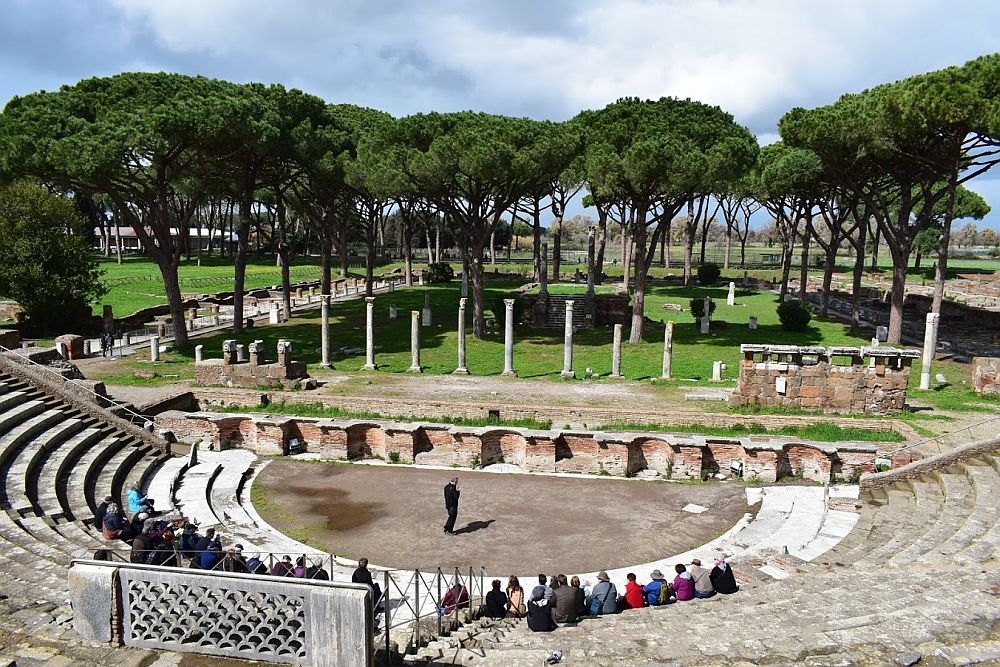 The ampitheater as seen from the top seats. The arc of the stone benches is visible at the bottom of the picture. A semi-circular stage area is at the bottom, and a man stands there, speaking to a small group of people seated on the lowest seats. Behind the stange area is a low wall, and behind that is a row of columns. Behind them is a large area with a number of trees.