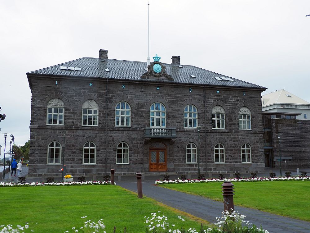 A dark brown stone building with two rows of windows across the front. The windows are all arched at the top. In the center of the facade is the entrance door, with a small balcony above it. 