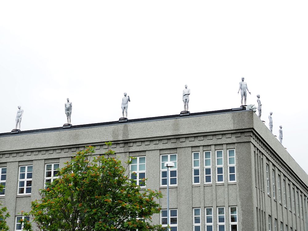 A blockish grey building, probably offices or a school, but right along the edge of the roof are a series of statues of men. All are full-body, white and stand facing out to the street. Each is posed slightly differently. Spotted during our free walking tour of Reykjavik.