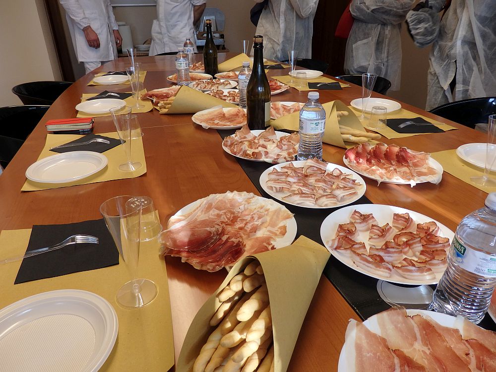A long table set with plastic plates around it. In the middle are a number of plates, each covered with cold cuts, each a bit different from each other. A bag in the foreground has bread sticks and here and there are bottles of wine and water.
