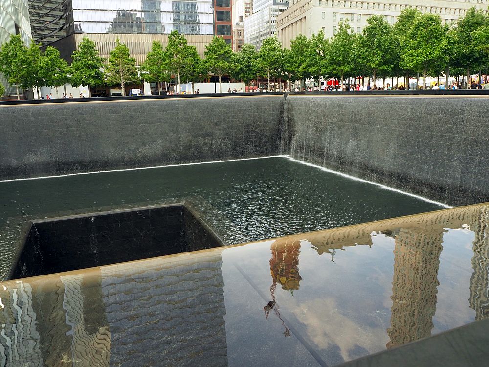 As described in the text. At the 9/11 memorila site, a square hole, with a square hole in its middle. Only two of the sides are visible, since the photo only shows one end of the fountain. The sides and bottom are dark gray. Water falls along both visible sides, but barely visible because it falls in a smooth curtain of water. People are visible on the other side, but extremely small, and only their heads and shoulders.