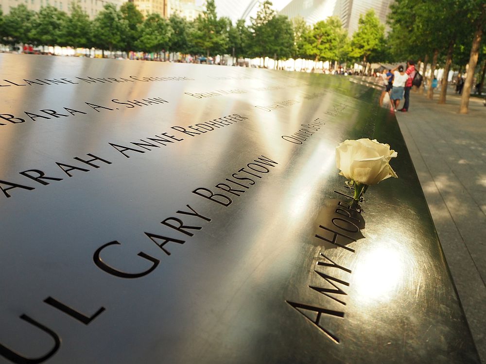 Looking along the bronze parapets of the 9/11 memorial site fountain. It has a flat surface that is tilted. A few names are partly visible: Sarah Anne Redheffer, Gary Bristow, Amy Hope ... her last name is obscured by a white rose that has been inserted into the first letter, L.