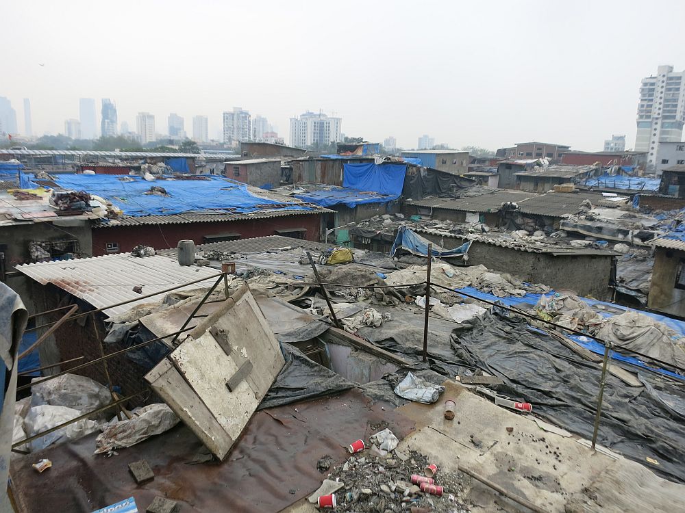 The rooftops look very poor: corrugated iron, but with tarps stretched over portions of them, with stones or other items to hold the tarps in place. In the distance, tall apartment buildings.