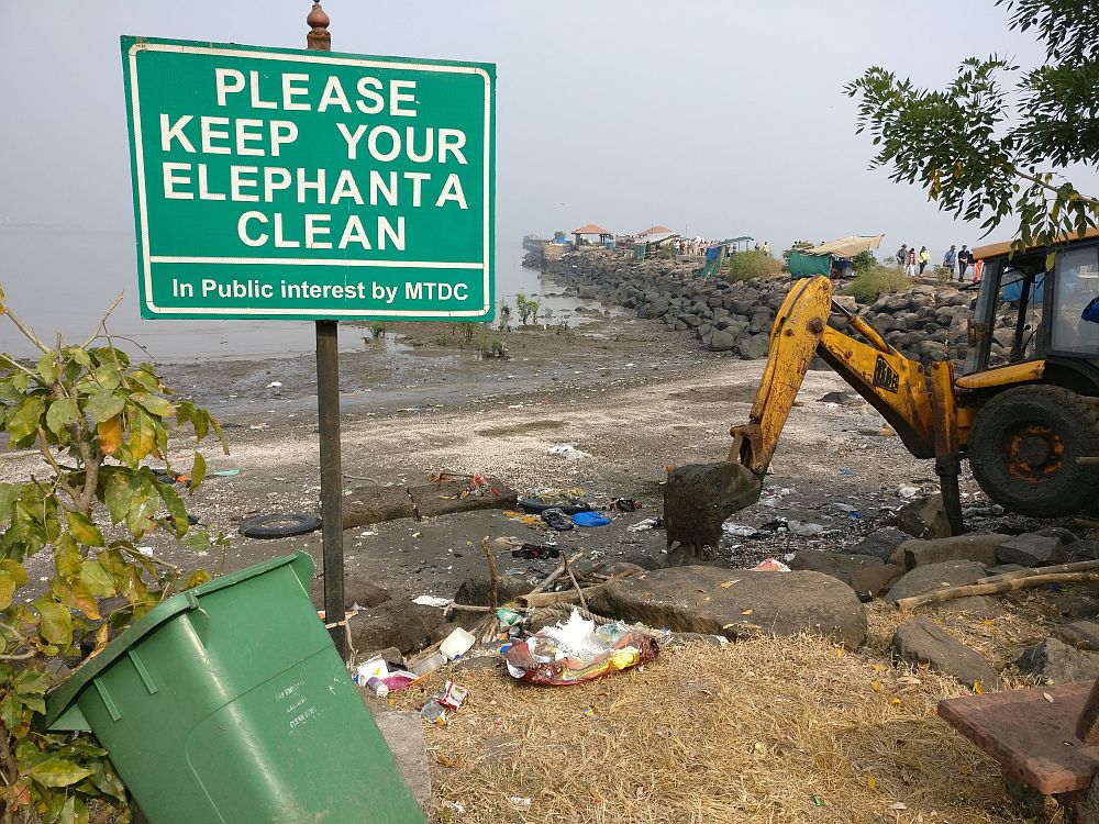 A view toward a beach with a jetty in the background. In the foreground, a green garbage bin lies on its side and the beach is strewn with garbage. How to be respectful when you travel.