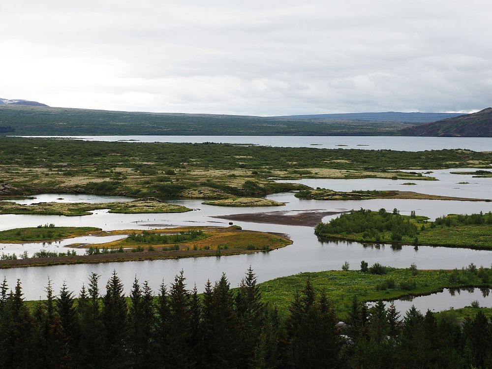 Big view over the valley, this time with water in the foreground: a wide river, with several low islands, and a bigger lake in the background. Beyond the lake: low hills. Gray sky above.