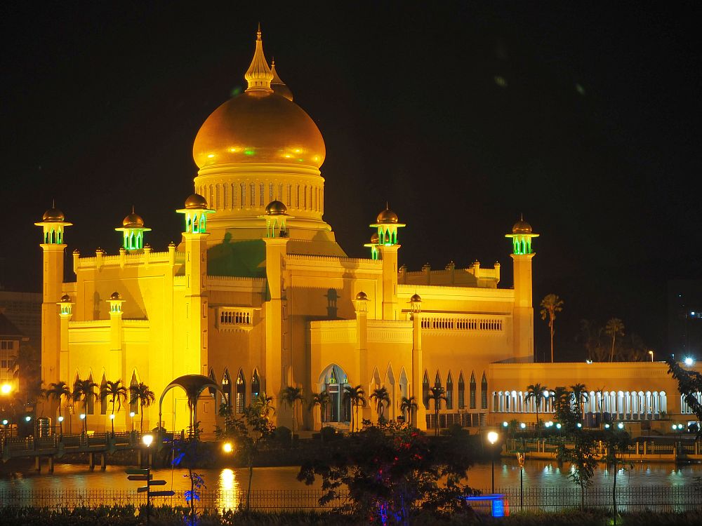 The mosque is pictured at night, light up from below. It has small turrets along its sides and a huge dome in the middle that is covered in gold. Around the building is a row of tall palm trees.