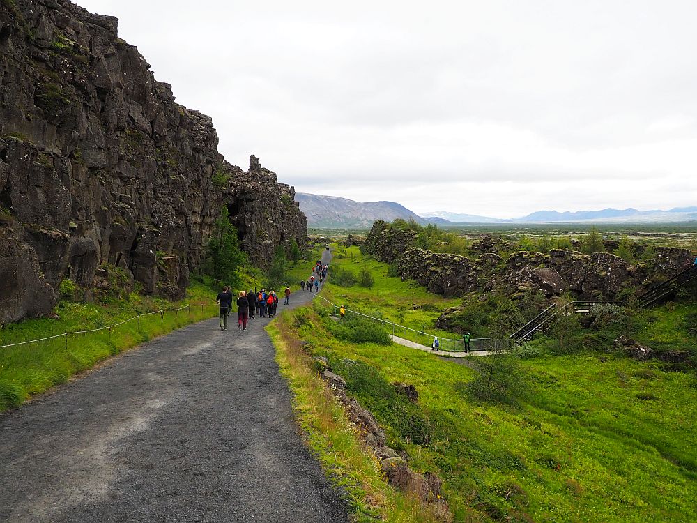 A wide grey gravel path stretches ahead, with a few people visible on it in the distance. On the left, a steep craggy cliff in black stone. On the right, a green slope down and then up to a lower rock wall. 