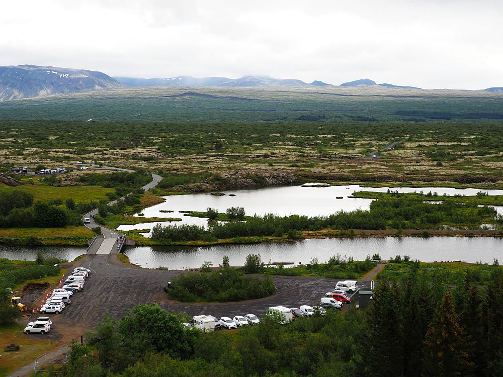 A big view over the valley, which has a flat bottom. On the horizon, some low mountains. In front of that, a flat expanse, some of it dirt/rock, some of it green. In the nearer distance, a river flows from across the picture from left to right and a lake is just behind it, separated by a narrow strip of land. In the near distance, well below camera level but near the bottom of the photo: two lines of cars are parked around a cleared dirt-floored area.