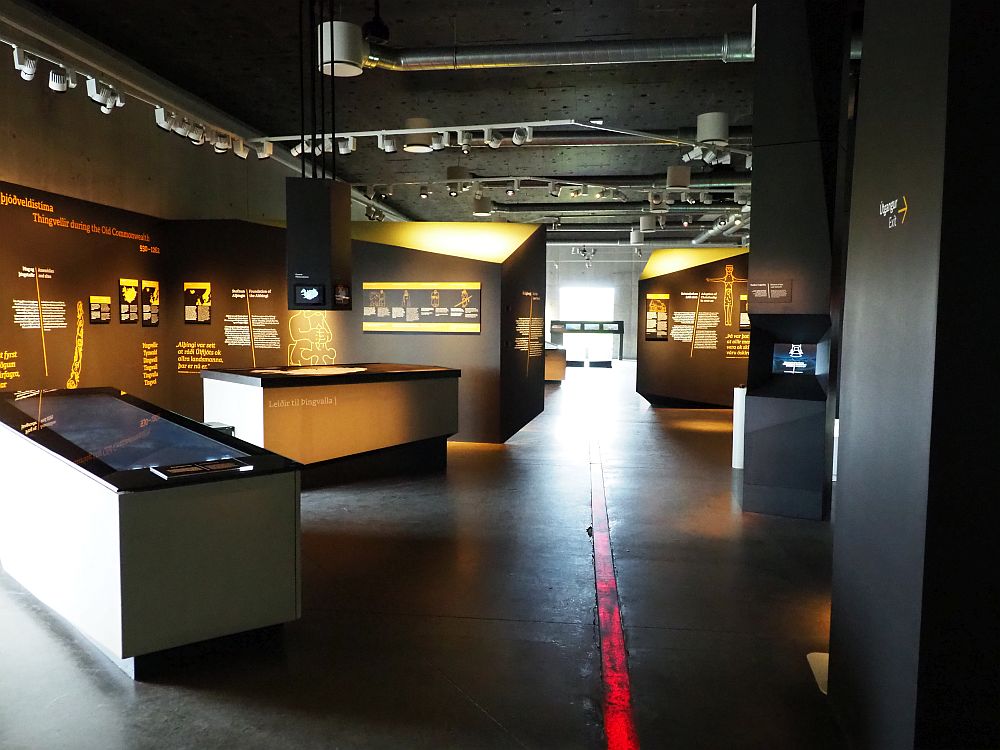 A long dark room with a black ceiling and floor, and a red lit-up line extending from the bottom of the photo to the end of the room. Lights expose various signs along the walls. Big blockish scrreen tables here and there, each with a slightly slanted top. 