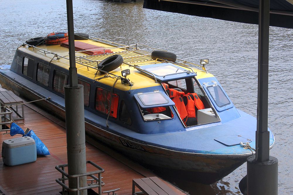 The boat is low to the water, painted blue, with a yellow roof with a luggage rack on it. Inside a few of the seats are visible, with bright orange life vests draped on them.