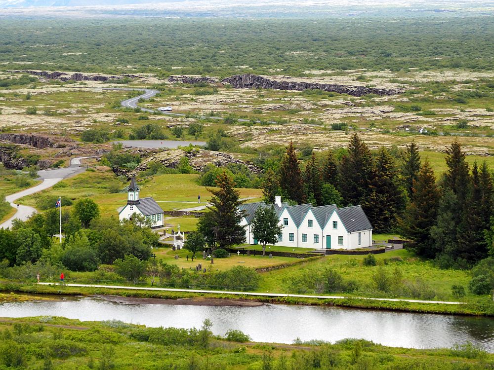 View into the valley with the church and prime minister's house in the center. In front of them, a small river. Behind them flat land stretches for a long way, some of it rocky, the rest green. About halfway to the top of the picture from the building is a crack in the ground with some rock walls showing.